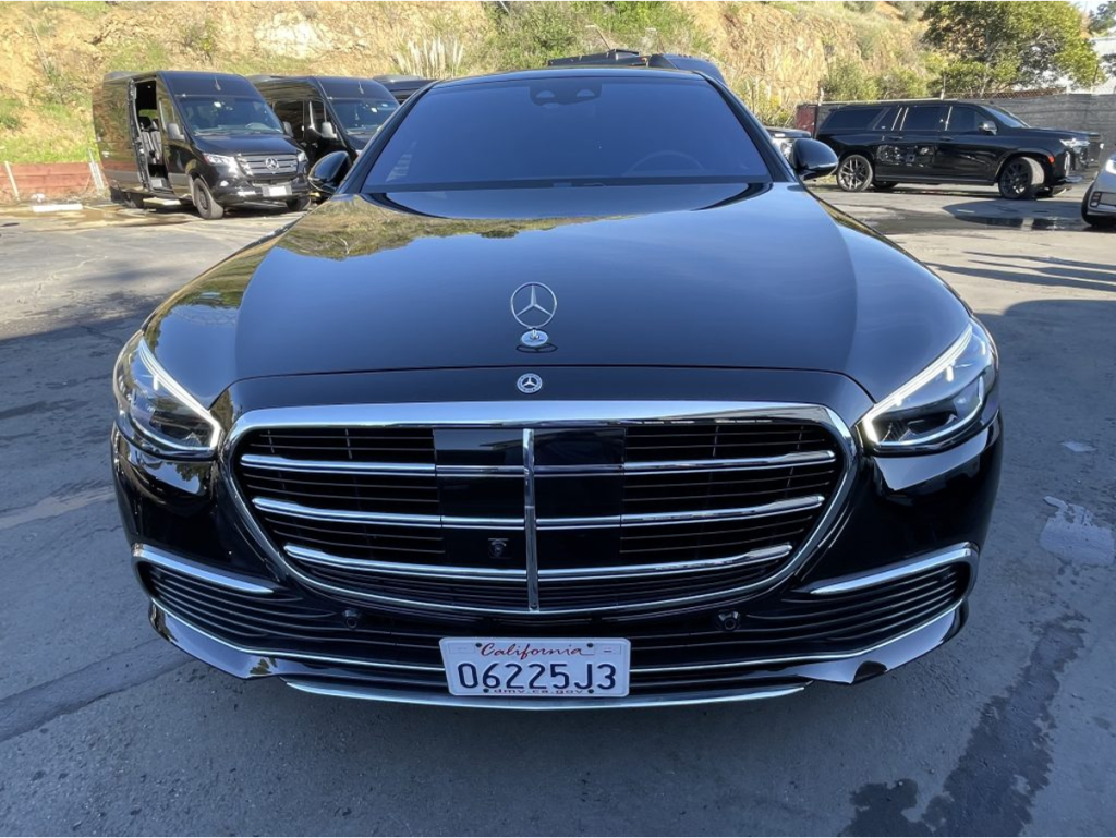 Black Mercedes S-Class sedan viewed from the front in a fleet parking lot, with a Mercedes Sprinter van and luxury SUV visible in the background