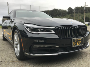 Black BMW 7 Series sedan with an SF VIP license plate parked in a lot with green hills and utility poles in the background under an overcast sky.