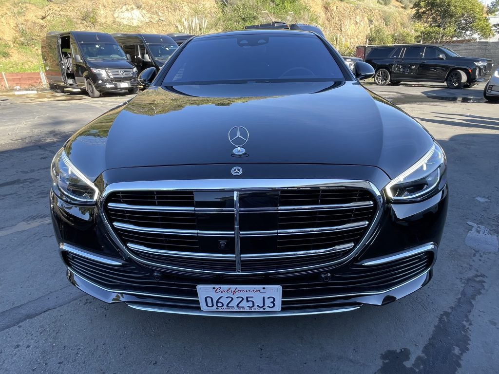 Black Mercedes S-Class sedan viewed from the front in a fleet parking lot, with a Mercedes Sprinter van and luxury SUV visible in the background.