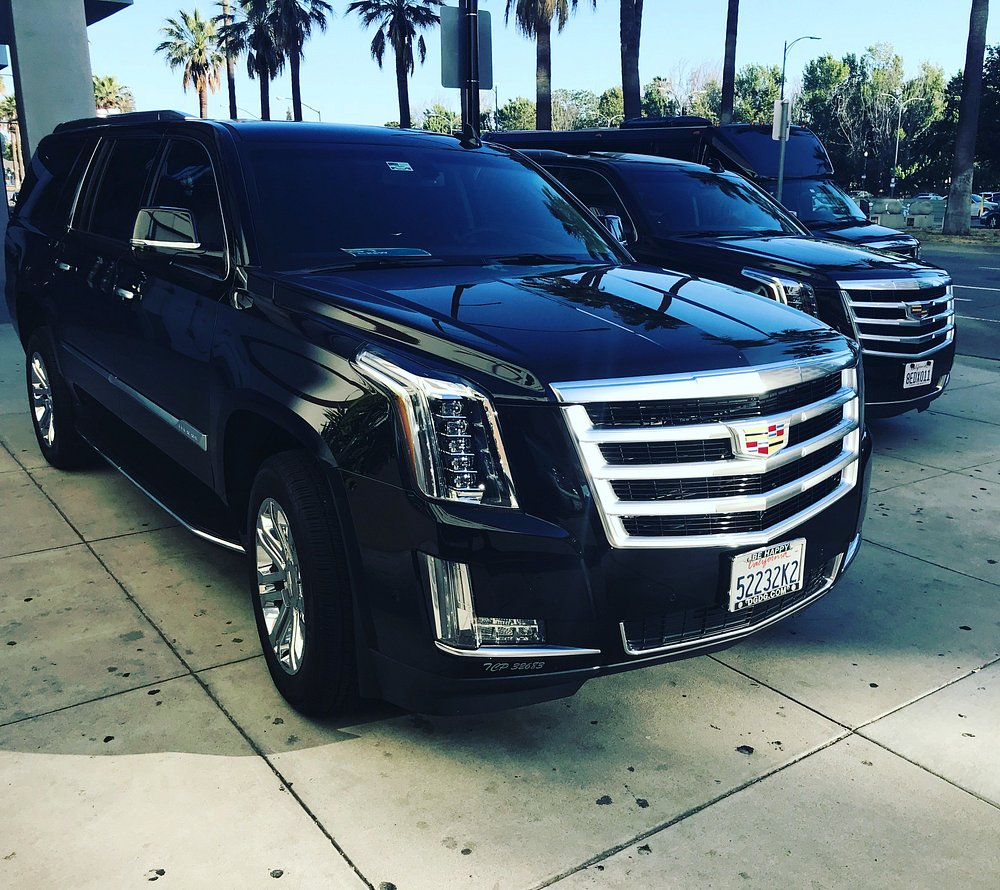 Two black Cadillac Escalade ESVs parked side by side on a palm tree-lined street on a sunny day.