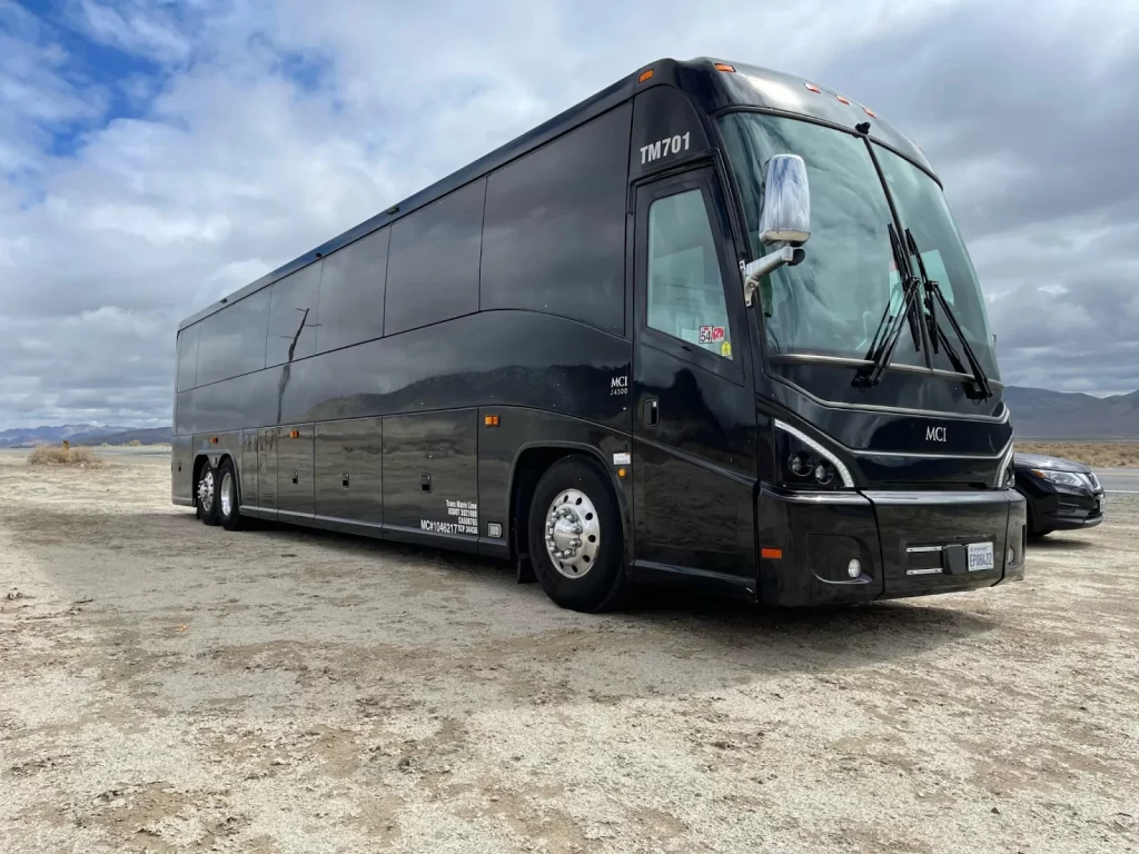 Black MCI motorcoach parked on a sandy lot under cloudy sky