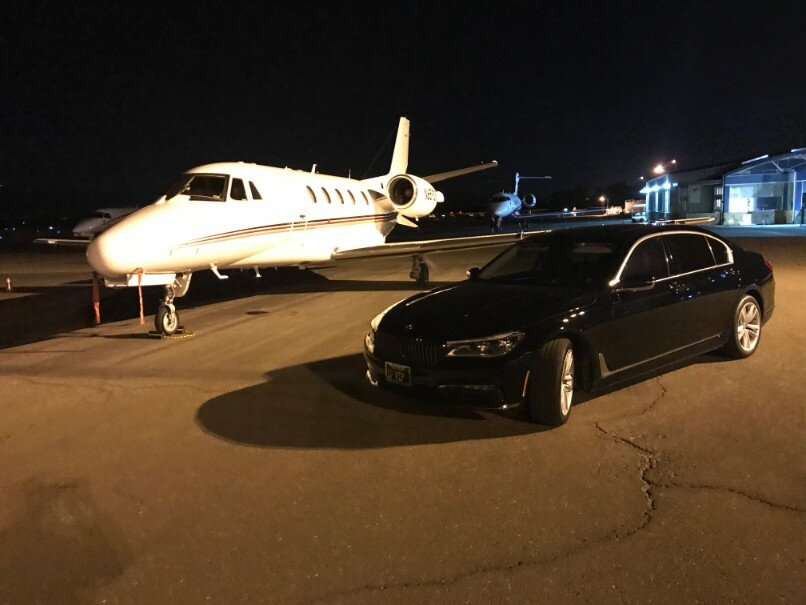 Black BMW sedan on a dark tarmac beside a parked jet, showing a late-night airport transfer.
